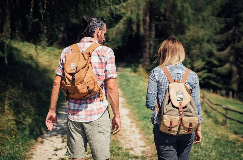 Zwei wandernde Personen mit Rucksäcken gehen auf einem Waldweg durch eine grüne Umgebung. Die Person vorne trägt ein kariertes Hemd, die andere Person eine Jacke mit Karo-Muster. | © Urlaub am Bauernhof Steiermark / Daniel Gollner 