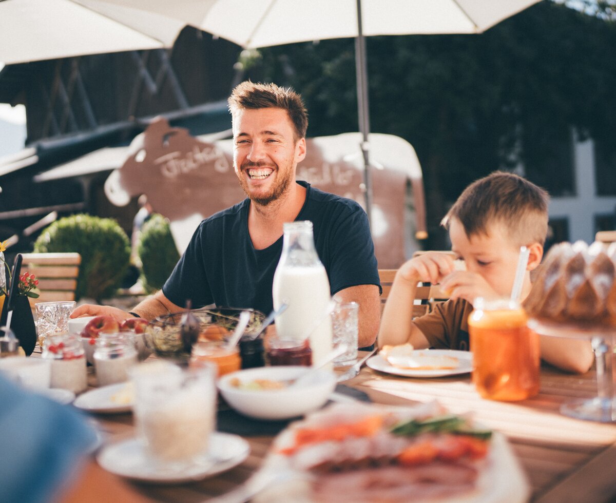 Familie beim Frühstück | © Urlaub am Bauernhof / Daniel Gollner Entspanntes Sommerwetter auf einer Terrasse. Eine Gruppe von Personen genießt ein Mittagessen mit Getränken und Kuchen. Im Vordergrund sieht man einen lächelnden Mann, der gemeinsam mit einem Kind am Tisch sitzt. | © Urlaub am Bauernhof / Daniel Gollner