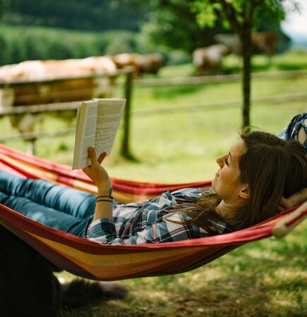 Eine Person entspannt in einer bunten Hängematte in einer grünen Landschaft mit Bäumen und Wiesen, liest konzentriert ein Buch. | © Urlaub am Bauernhof / Daniel Gollner 