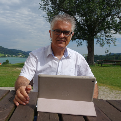 Ein Mann mittleren Alters in weißem Hemd und Brille sitzt an einem Holztisch im Freien. Im Hintergrund ist eine grüne Landschaft mit Bäumen zu sehen.